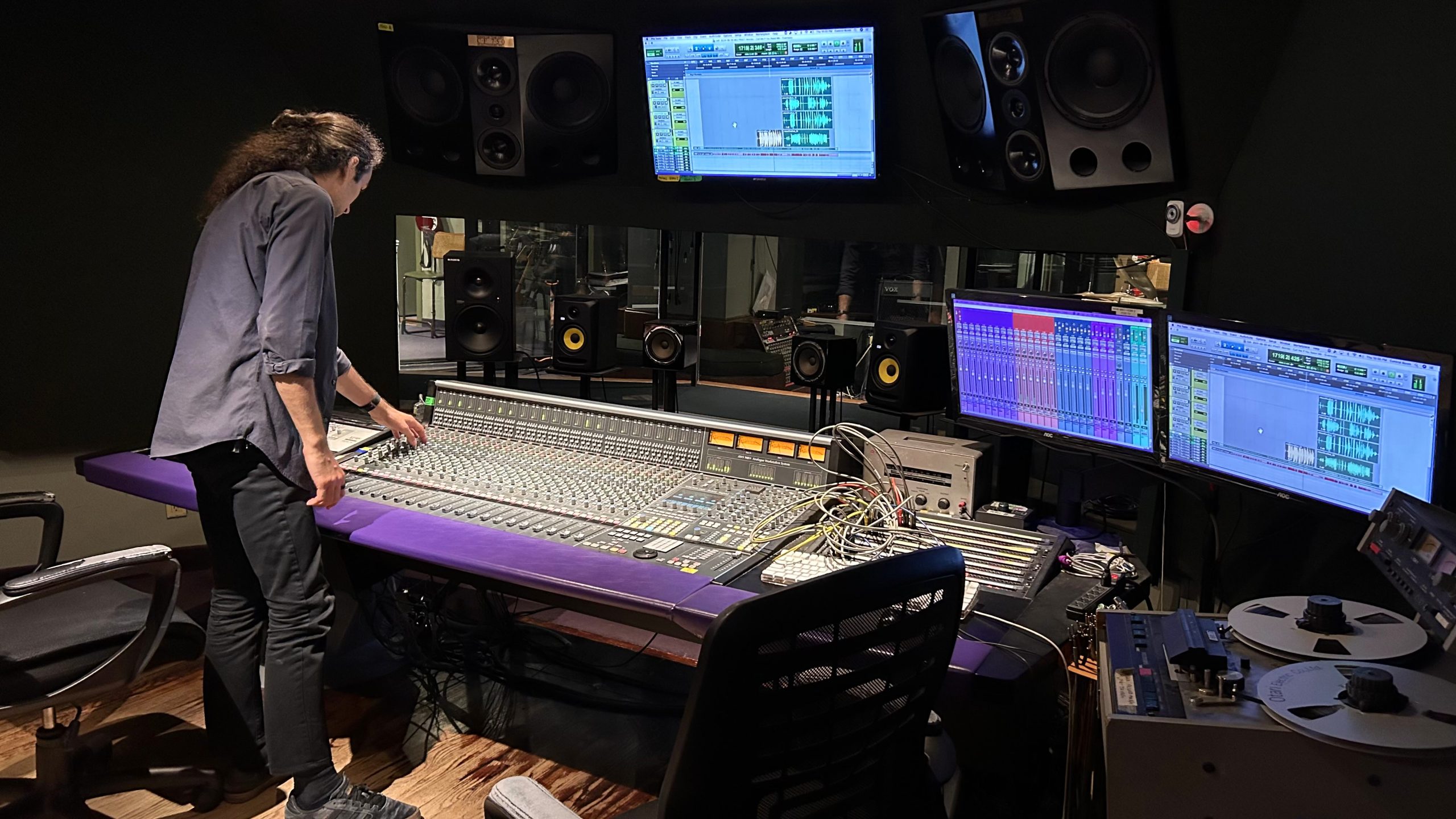 Photo of Sam in control room, standing at mixing console. Perspective is from back of control room.