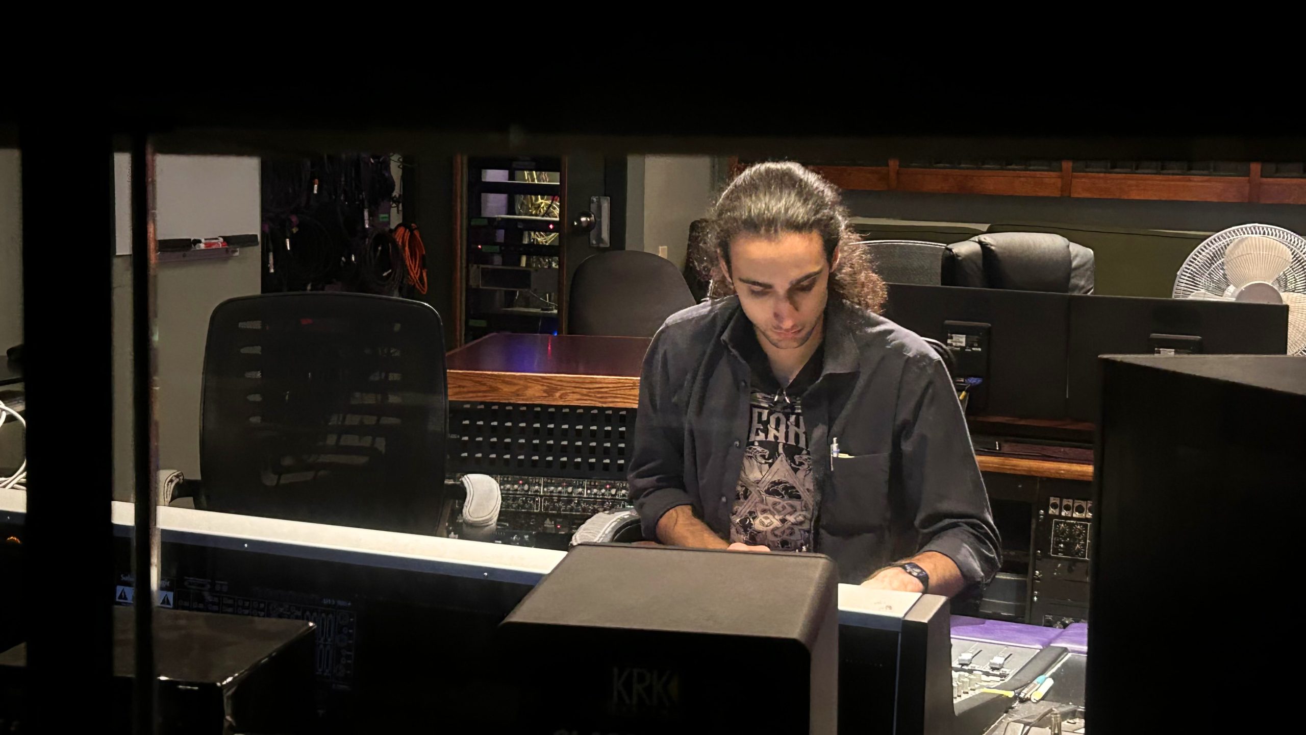 Photo of Sam in control room, sitting at recording console. Perspective is from outside of control room, looking in.